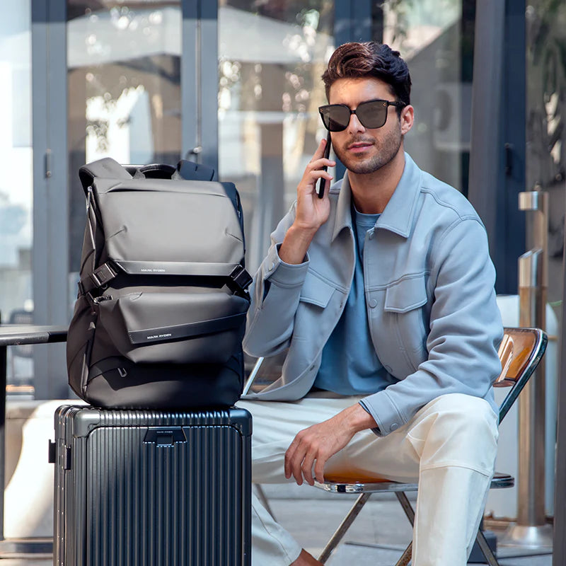 Mark Ryden 2859 black roll-top backpack placed on a suitcase next to a man sitting outdoors and talking on a phone, illustrating a modern travel and business lifestyle setting
